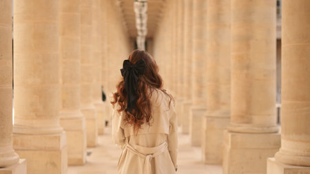 Woman in trench coat with black bow walks through colonnade