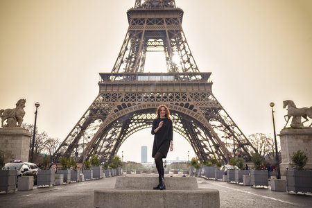 Cheerful young woman poses with arms spread in front of the Eiffel Tower on an overcast day in Paris.の写真素材