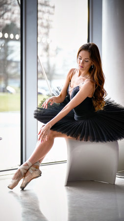 Graceful female ballet dancer in black tutu and pointe shoes seated by the window, posing with elegance and calm expressionの写真素材