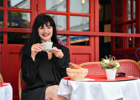 Happy woman enjoying coffee at a charming Parisian cafe with a croissant and flowers on the table, framed by red chairs and a bright, authentic backdrop.の写真素材