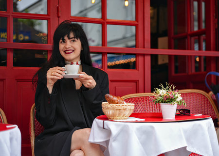 Smiling woman enjoying coffee at a Parisian street cafe with a croissant and flowers on a red table, captured in a warm and joyful lifestyle moment.の写真素材