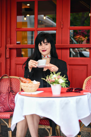 Charming woman holding a coffee cup at a classic Paris cafe table with croissant, flowers, and red details, smiling softly in a cozy outdoor setting.の写真素材