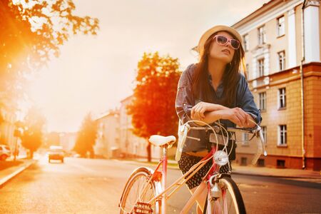 portrait of a beautiful young girl in a hat with a bicycle on city background in the sunlight outdoorの写真素材