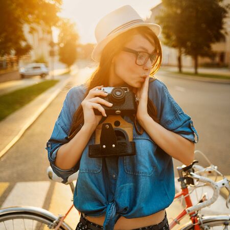 stylish woman in hat standing near bike in morning sunshine with photo camera outdoorの写真素材