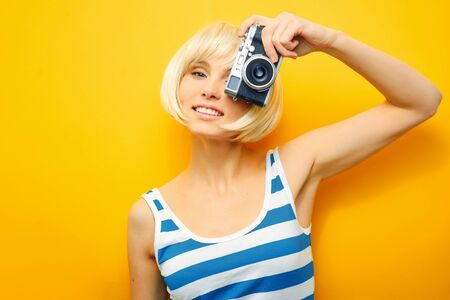 Portrait of a smilling girl with a camera in hand on a yellow background. Isolated studio. blonde wigの写真素材