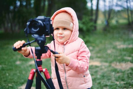 Child with photo camera in a city park. Girl make a video and photo for the Internet, record a video blog for camera. pink jacket and hatの写真素材