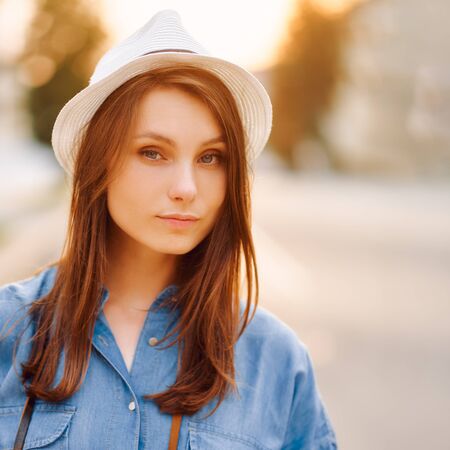 portrait of girl in a white hat and a blue denim shirt on a background of a sunset cityの写真素材