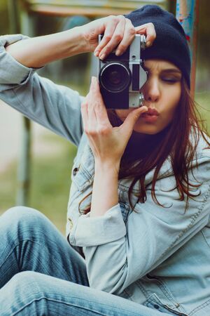 portrait of a beautiful hipster girl in a cap and a denim jacket with a camera taking photosの写真素材