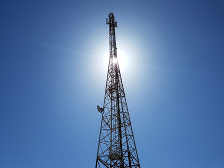 low angle view of cellular or mobile network tower against the sun in the blue sky.