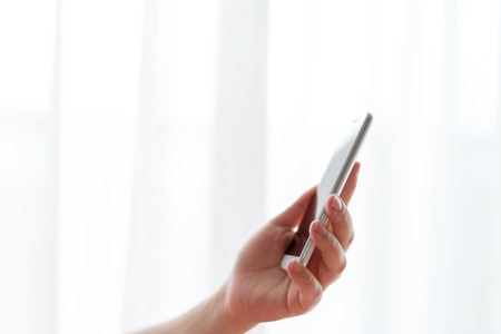 Woman sitting in the chair and holding a mobile phone in front of the window with curtainsの写真素材