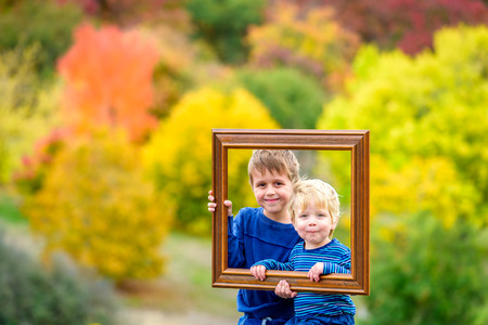 Two smiling kids in autumn park looking into camera through wooden frameの写真素材