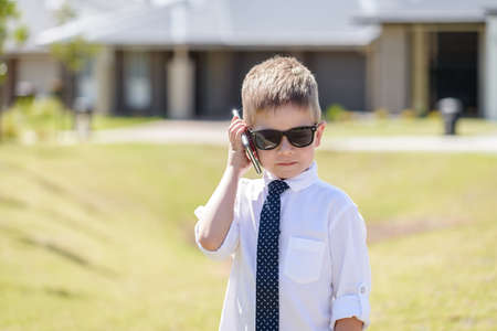 Cute Australian boy wearing business attire with sunglasses while using mobile phone on a bright summer dayの写真素材