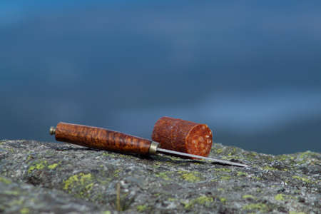 Knife cutting sausage in the mountain with blurry mountain landscape in background. Outdoor food.の写真素材