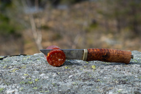 Knife cutting sausage in the mountain with blurry mountain landscape in background. Outdoor food.