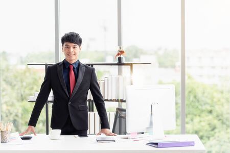Portrait of a confident happy young businessman wearing a suit standing at a desk in a modern office.の写真素材