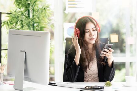 One beautiful working woman sitting at the office desk in the office and wearing headphones listening to music from a smartphone during the break. She smiled and enjoyed being relaxed.の写真素材