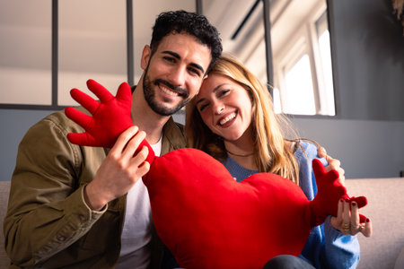 Young couple celebrating Valentines day, smiling and hugging a heart looking at cameraの写真素材