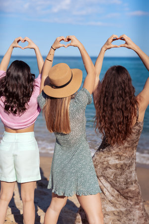 Three girls standing in front of the sea making heart shapes with their handsの写真素材
