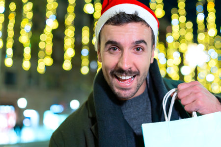 Man Santa hat smiling holding shopping bags festive city street night lights
