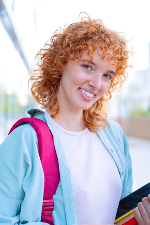 Cheerful red-haired student holding a red folder in a bright outdoor setting