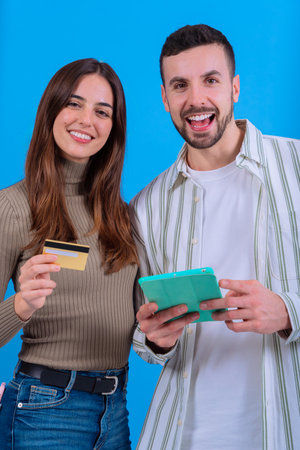 Happy couple holding a credit card and tablet against a blue background.Vertical