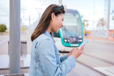 Smiling Young Woman Using Smartphone While Waiting for Tram at Stationの写真素材