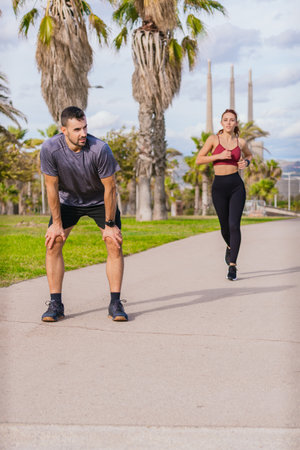 Tired man resting after jogging while woman continues running in a park.Vertical