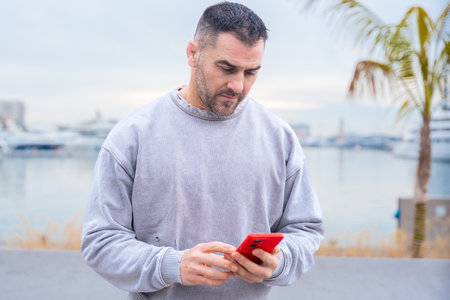 Man typing on red smartphone while sitting outdoors near palm trees and portの写真素材