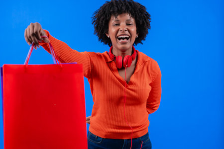 Happy young woman holding a red shopping bag, smiling against blue background.の写真素材