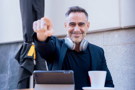 Smiling man pointing at camera while working with tablet and coffee in outdoor cafeの写真素材