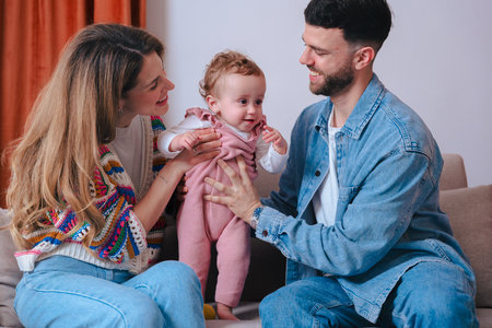 Young parents smiling while helping baby girl stand on sofa during family time