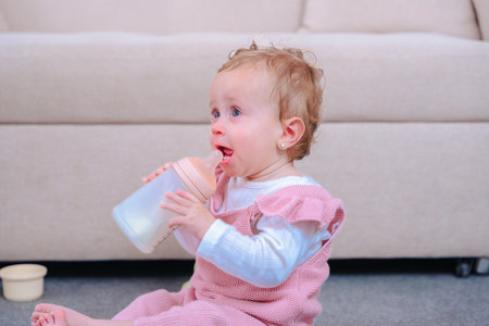 image of baby girl drinking milk from bottle while sitting on carpeted floor