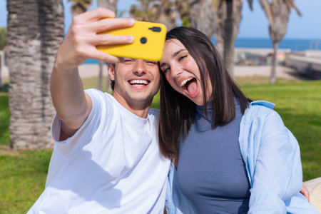 Playful young couple taking selfie with yellow phone in seaside palm lined park