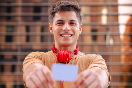 Smiling young man with red headphones showing card in urban environment