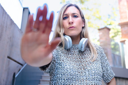 Woman gesturing stop with hand towards camera while wearing headphones outdoors