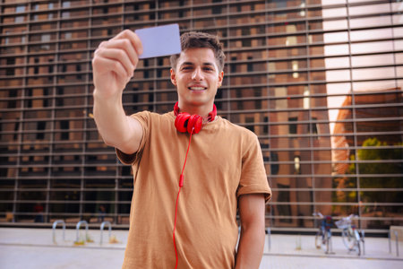 Smiling young man with red headphones showing card in urban environmentの写真素材