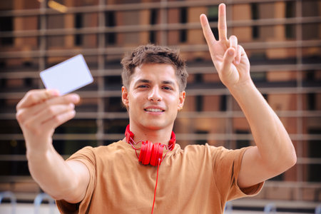 Young man holding blank white card showing peace sign outdoors with red headphones