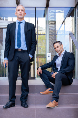 Two businessmen in suits posing outside modern office building, serious look.の写真素材