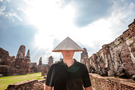 Portrait of man in chinese hat standing near the ancient ruins at Wat Mahathat in Ayutthaya historical park at Ayutthaya,Thailand while sunbeams shining through the cloudsの写真素材