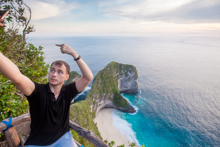 Man doing funny selfie on the smartphone with incredible green rocks shaped like dinosaur head with...