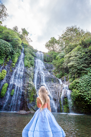 Young woman with long hair in skirt standing back in front of Banyumala twin waterfalls with cascades among green tropical trees and plants on the north of Bali island, Indonesiaの写真素材