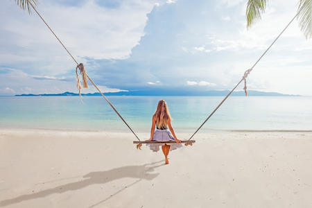 Young girl with long hair sitting back on the swing at the beach near the tropical sea with island on the backgroundの写真素材