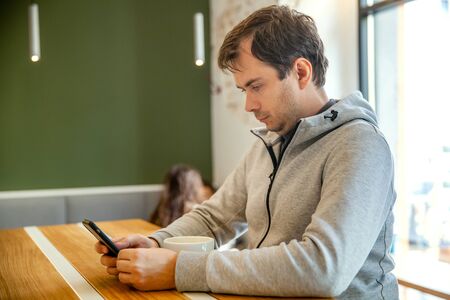 Serious man sitting in cafe with cup of coffee, working with smartphone