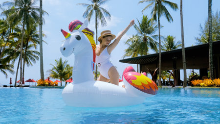 Traveling in Thailand. Happy smiling woman fun, dancing on big inflatable unicorn toy mattress in swimming pool and celebrating summer vacation in tropical resort with palm trees and seaの写真素材