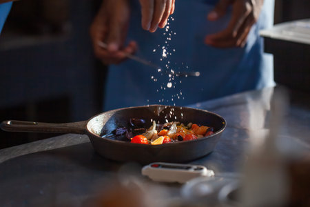 Chef cook preparing vegetables in a luxury restaurant or hotel kitchen. Man hands sprinkle salt on roasted vegetables in pan. Professional cook in uniform add some species to dishの写真素材