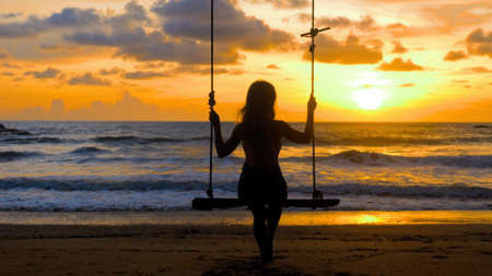 Young woman swinging sitting on wooden swing with ropes on sandy beach, sea sunset background. Girl silhouette enjoy outdoors freedom, summer. Travel, tourist, holidays, vacations concept. Back viewの写真素材