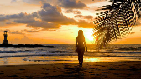 Attractive girl walking on sandy beach near palm tree branch with sea, sunset sun, lighthouse on background. Young woman silhouette enjoy summer holidays. Travel, tourist, vacations outdoors conceptsの写真素材