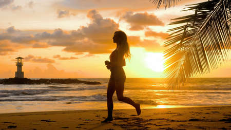 Young woman exercising running barefoot on sea beach with sunset ocean background. Summer seascape, healthy active lifestyle, outdoor jogging sports concept. Female athlete silhouette cardio trainingの写真素材