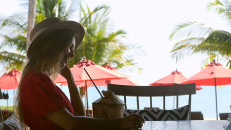 Side view of travel woman sitting in outdoor cafe with young fresh green coconut, enjoy summer and sea view. Female traveler on vacation in modern resortの写真素材