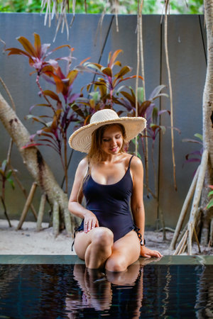 Travel woman in straw hat and black swimsuit sitting on pool edgeの写真素材